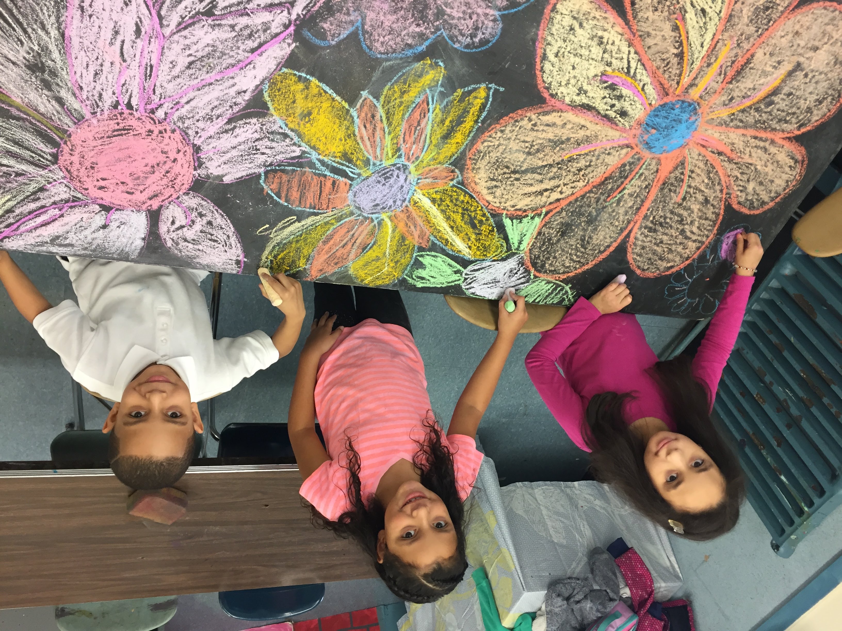 Three students collaborating on a large floral chalk drawing across a classroom table