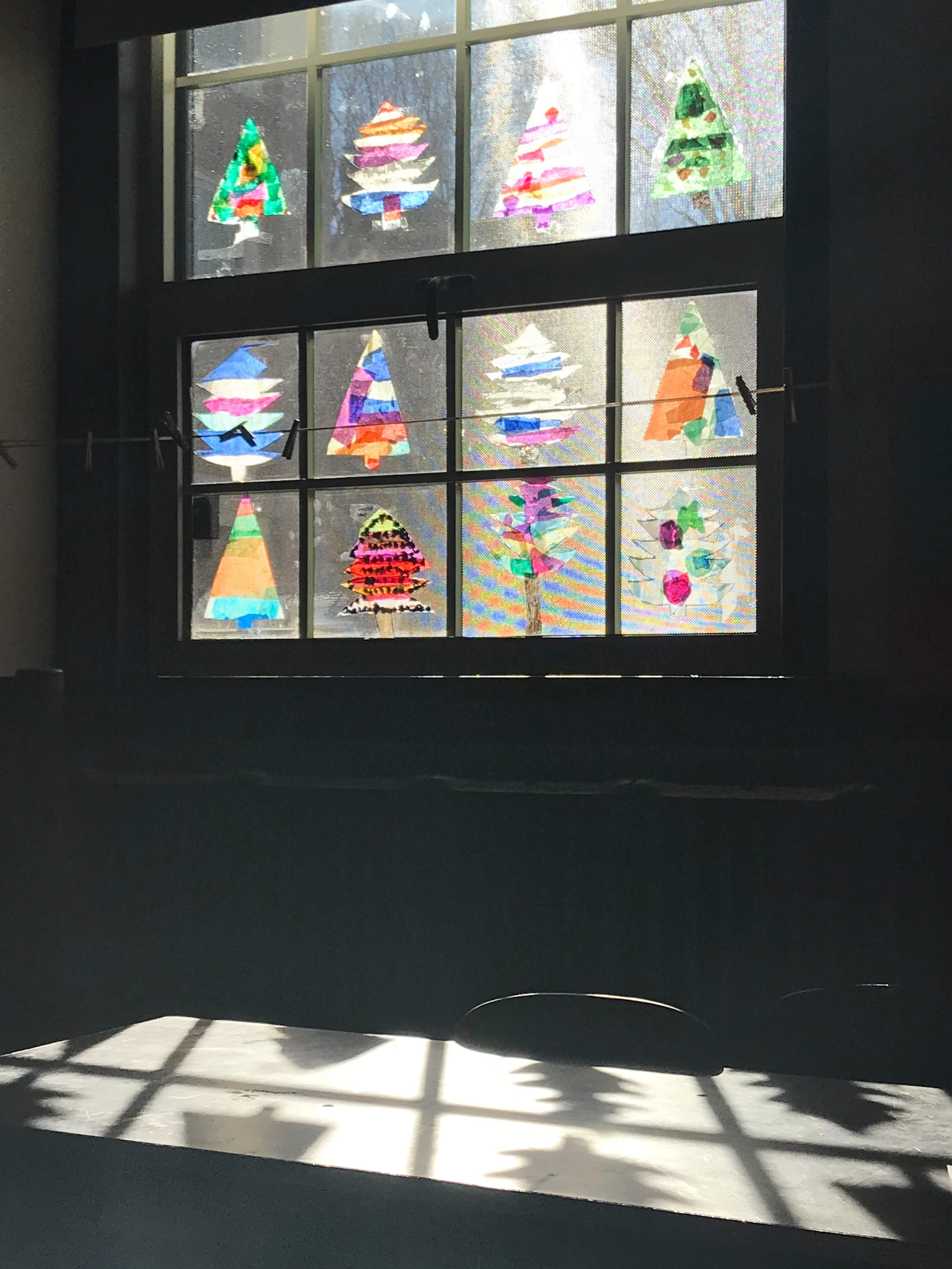 Colorful student tree collages installed in classroom windows, casting reflected light and shadows across a table